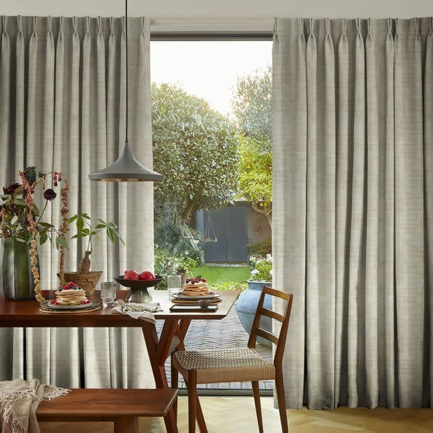 Pleated, floor-length grey curtains, drawn aside to frame a central opening and admit soft daylight; a wooden dining table set for breakfast beneath a pendant lamp, overlooking a sunlit garden.