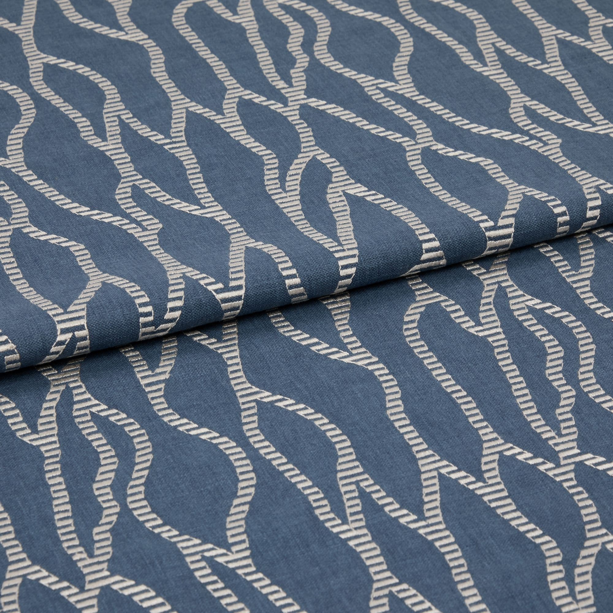 Blue woven fabric with cream embroidered wavy rope pattern, folded over itself, shown in a close-up studio view with soft, even lighting.
