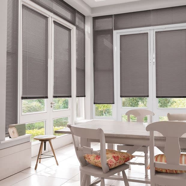 Gray horizontal slatted blinds, fitted inside tall white-framed windows and a glass door, partially lowered and filtering daylight, over a bright dining nook with white table and garden view.