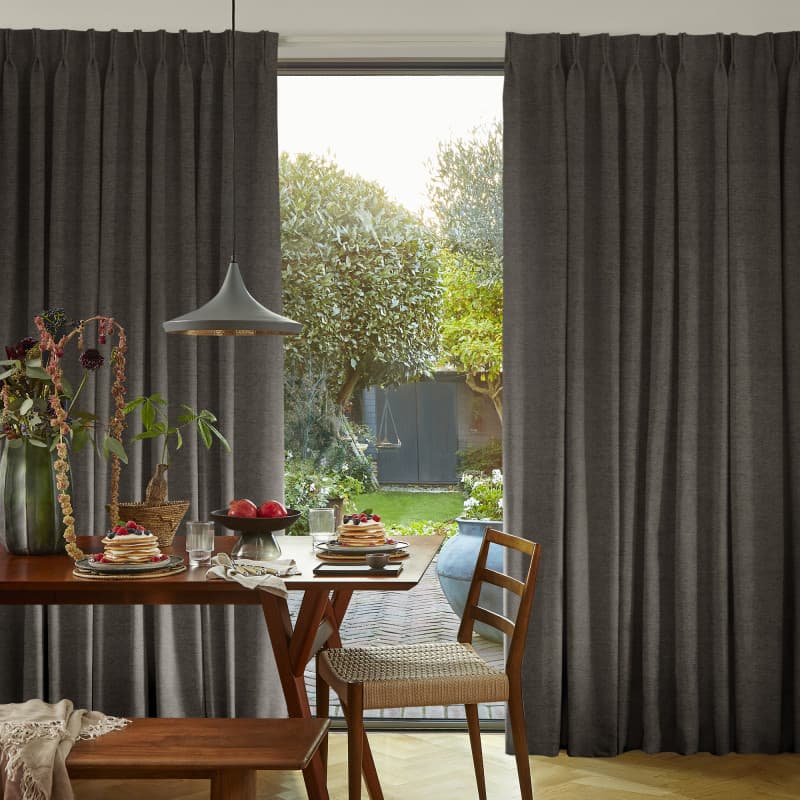Floor-length pleated dark-gray curtains, partly drawn to frame a central opening, letting daylight into a dining room with wooden table set for breakfast, a chair, and a sunlit garden beyond.