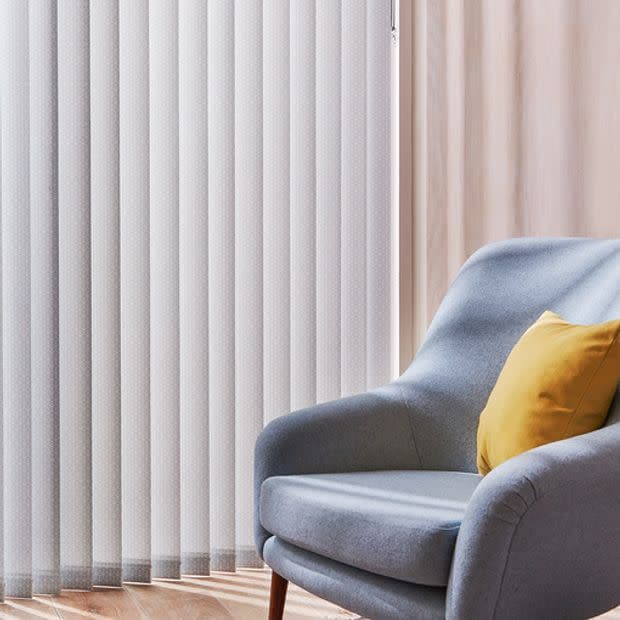 Vertical fabric blinds covering a floor-to-ceiling window, closed, softly diffusing daylight; beside them a light grey upholstered armchair with a yellow cushion in a lit living room with wooden floor.