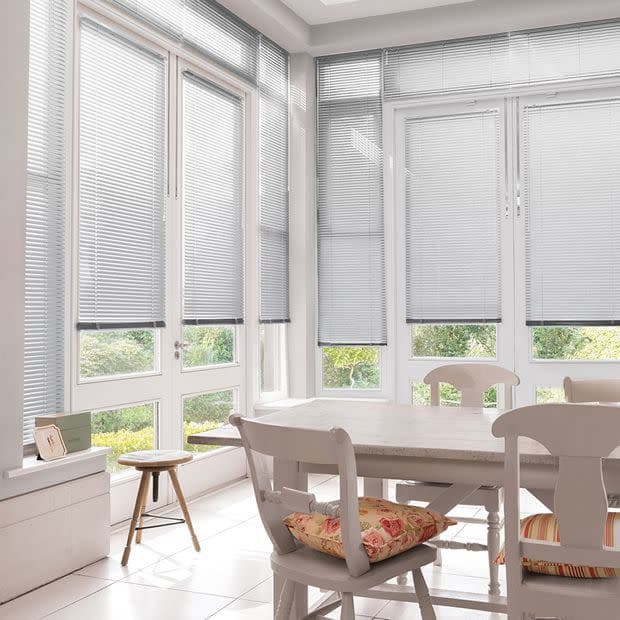 Horizontal aluminum Venetian blinds on tall bay windows, lowered to mid-height and filtering soft daylight, in a bright dining nook with a white table, chairs, cushions, tiled floor, and garden view.