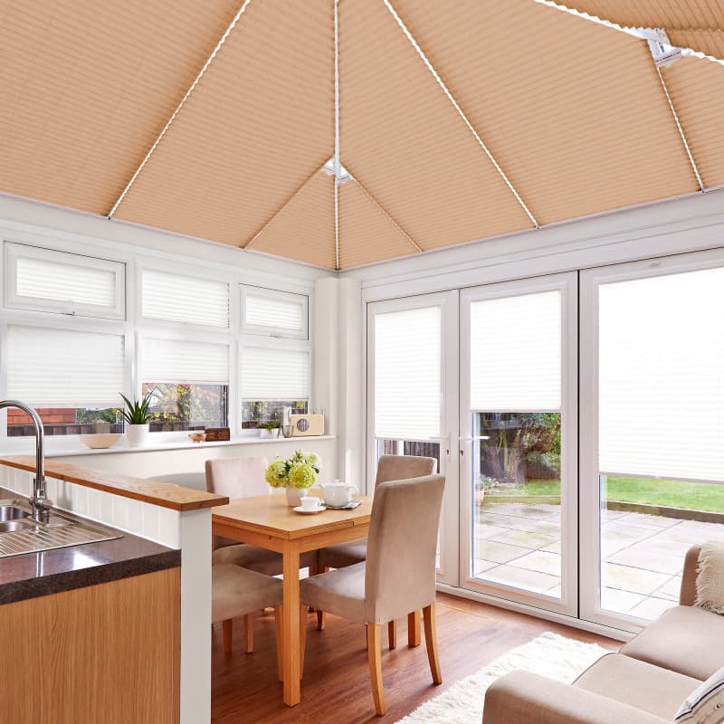 Beige pleated roof blinds and white pleated window/door blinds, fully lowered, filtering soft daylight; in a bright conservatory dining area with wooden table, upholstered chairs, sink and garden patio view.