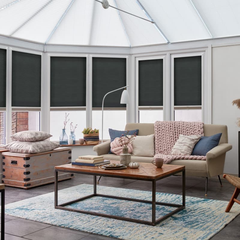 Dark grey cellular pleated blinds covering bay conservatory windows, lowered to midâ€‘height and blocking light; sunroom with beige sofa, chunky pink throw, wooden coffee table on blue rug.