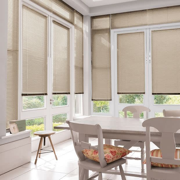 Beige horizontal Venetian blinds covering tall corner doors and windows, narrow slats lowered to the sill filtering daylight; bright sunlit dining nook with white table, chairs, tiled floor, garden view.