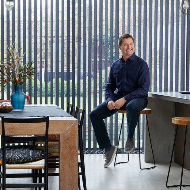 Floor-to-ceiling vertical blinds with narrow dark slats, mostly closed yet permitting garden views and daylight; modern kitchen/dining interior with wooden table, vase, man sitting on barstool smiling.