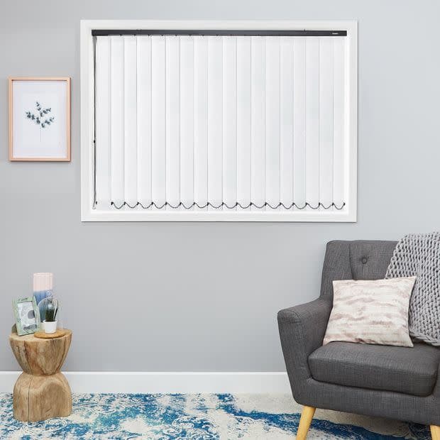 White vertical blinds covering a central window, closed with visible headrail and linked bottom chains, diffusing daylight; in a grey-walled living room with a grey armchair and blue rug.