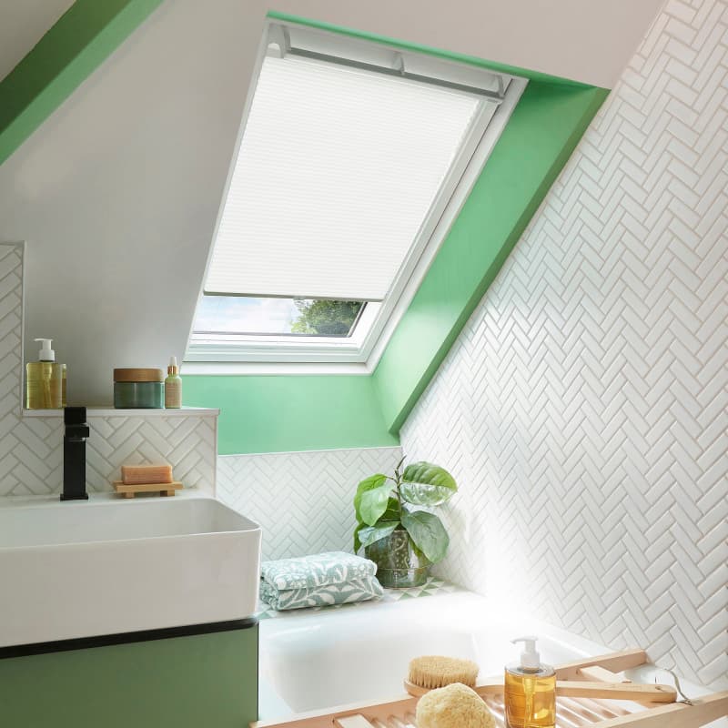 White pleated skylight blind lowered over a slanted roof window, filtering daylight; set in a green-trimmed attic bathroom with white herringbone tiles, sink, bathtub, potted plant, and bath tray.