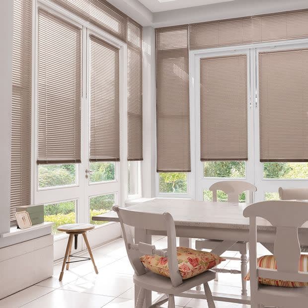 Beige horizontal Venetian blinds covering tall bay windows and doors, mostly lowered and filtering daylight; sunlit dining nook with white table and chairs, floral cushions, tiled floor, garden view.