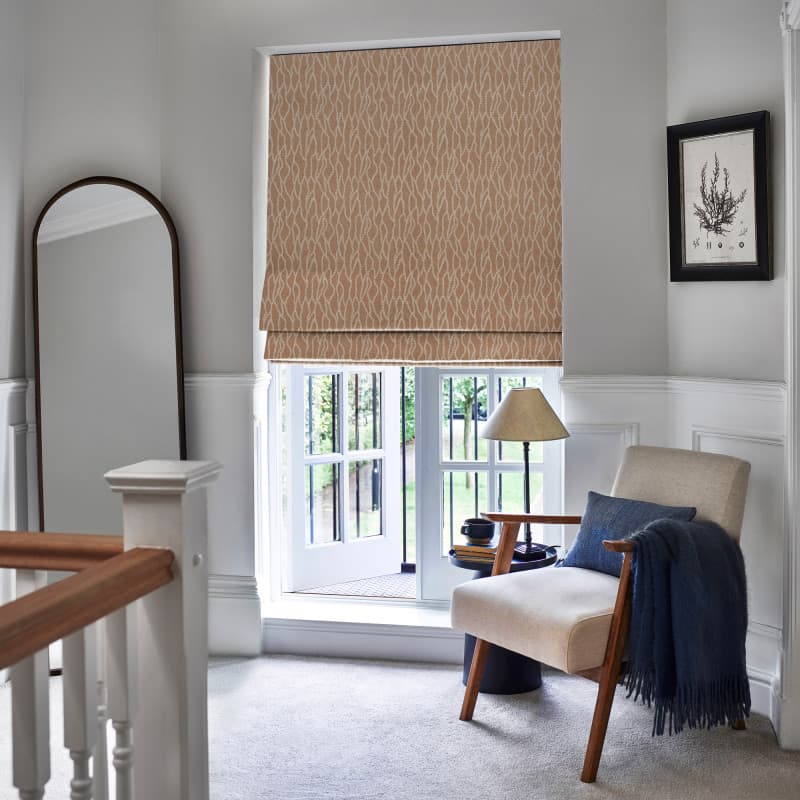 Patterned beige Roman blind, lowered and slightly folded at the bottom, filtering daylight over a ground-floor casement window opening to a garden, in a lit landing with armchair, lamp, mirror.