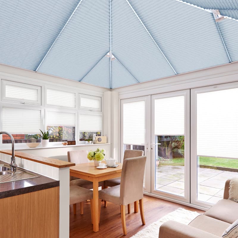 Blue pleated roof blinds fitted to the pitched conservatory ceiling, with white pleated window and door blinds lowered, filtering soft daylight over a dining area, kitchen sink, and garden view.