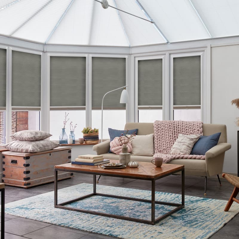 Gray pleated cellular blinds, lowered inside white-framed conservatory windows and filtering sunlight; situated above a beige sofa and wooden coffee table in a softly lit, pastel-accented living room.