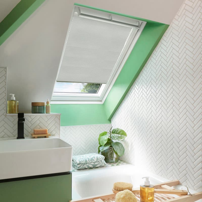 White pleated skylight blind, partially lowered over a slanted roof window, filtering daylight; set above a bathtub in a bright bathroom with green trim, white herringbone tiles, sink and plant.