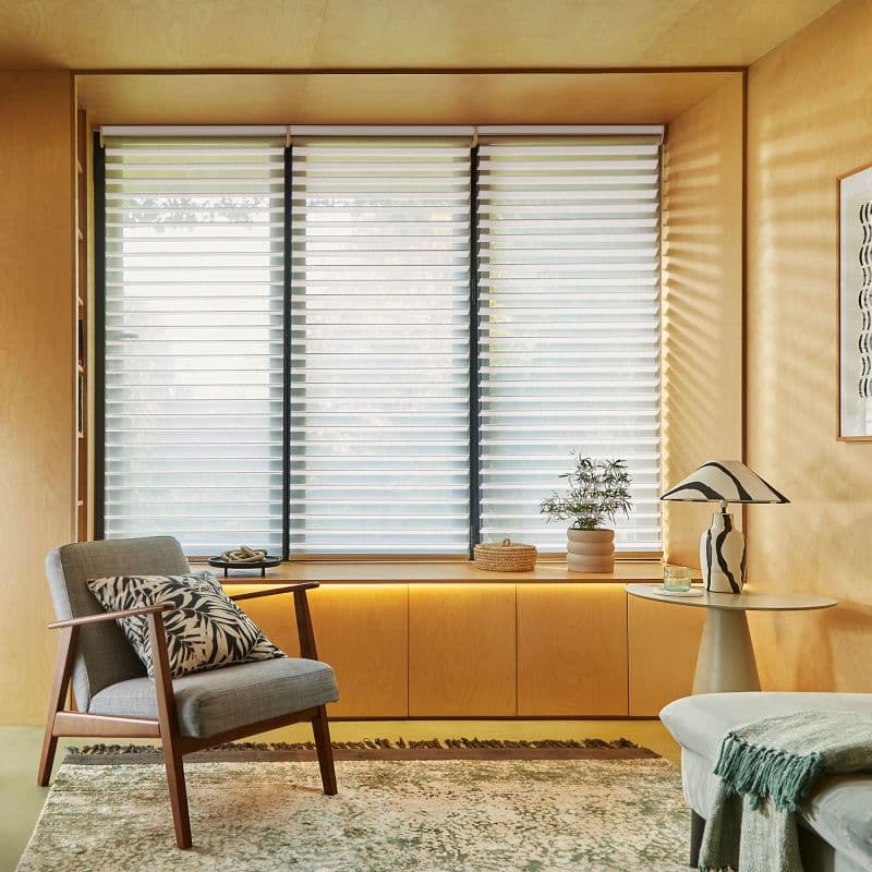 White horizontal slatted blinds covering a three-panel window, filtering soft daylight through semi-closed slats; in a warm wood-paneled sitting nook with armchair, potted plant, table lamp, and rug.