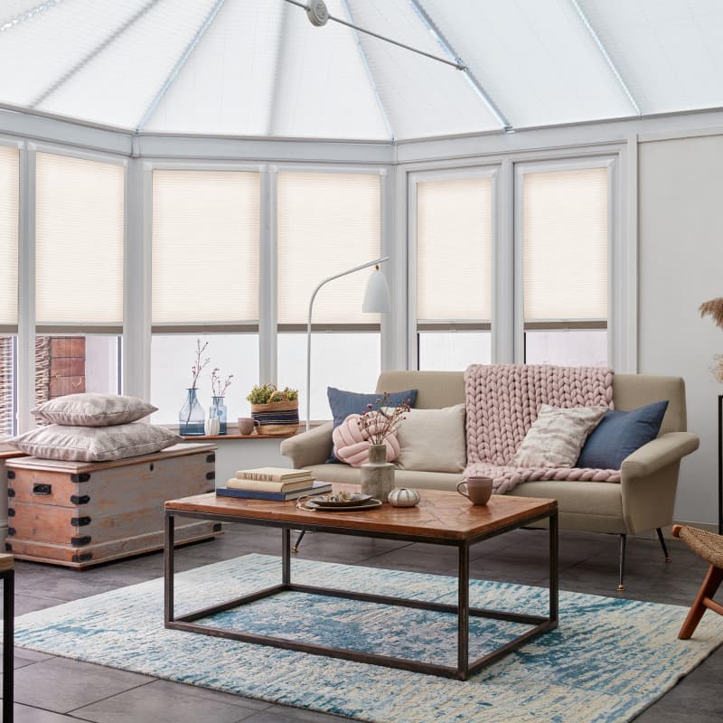 Pleated cellular blinds, fully lowered on bay windows with lightâ€‘filtering opaque fabric and narrow horizontal pleats; filtering soft daylight into a conservatoryâ€‘style living room with sofa, coffee table, rug.