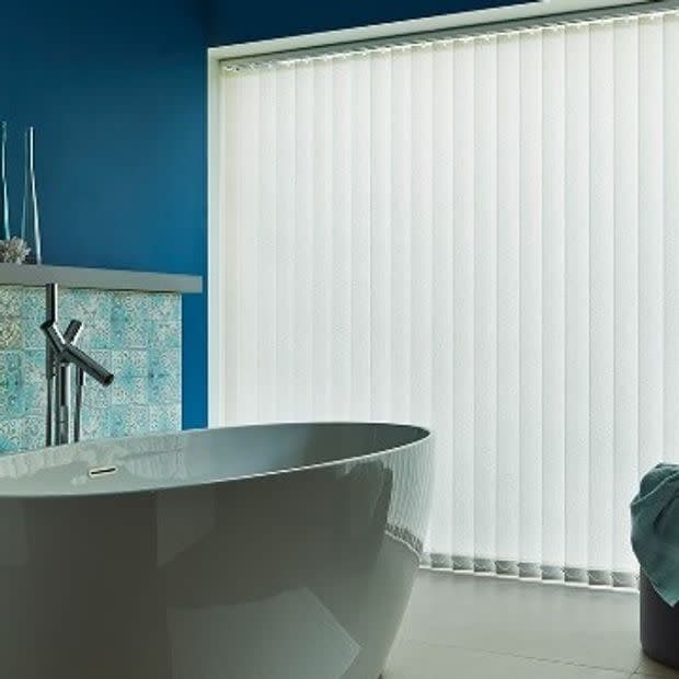 Vertical blinds covering a floor-to-ceiling window, closed and filtering soft daylight; in a modern bathroom with a white freestanding tub, blue accent wall and patterned tile backsplash.