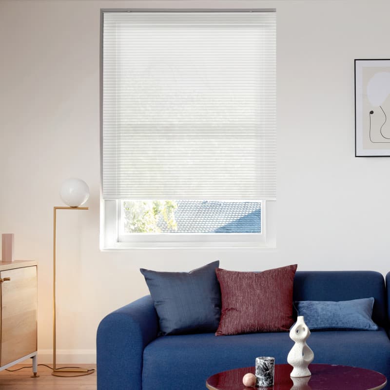 White pleated blind, fully lowered with horizontal texture, filtering soft daylight; living-room window above a blue sofa with cushions, gold floor lamp, wooden sideboard, framed abstract art and rooftop/trees view.