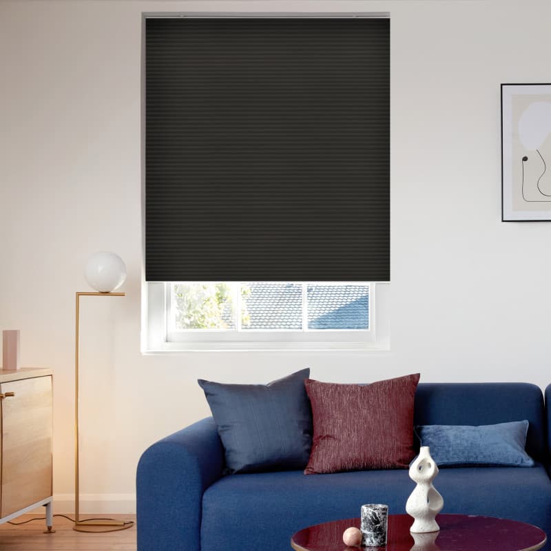 Black pleated cellular blind, fully lowered and blocking light with a narrow bottom gap revealing roof and trees; over a living-room window above a navy sofa, lamp, and coffee table.