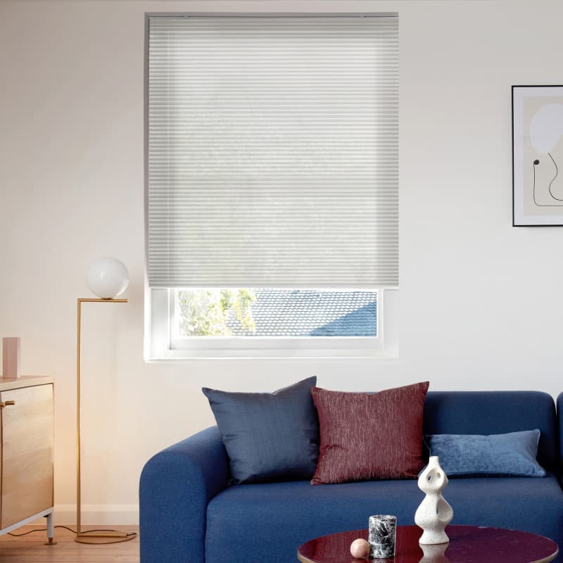 White cellular pleated blind lowered across window, translucent and filtering soft daylight; small modern living room with blue sofa, textured cushions, brass floor lamp, coffee table and framed abstract art.