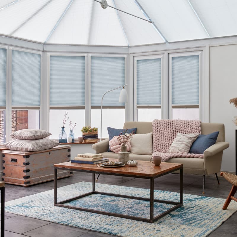Pale-blue pleated blinds fitted inside bay windows and the conservatory roof, lowered and gently diffusing daylight; cozy sunroom living area with sofa, chunky pink throw, wooden coffee table, blue rug.