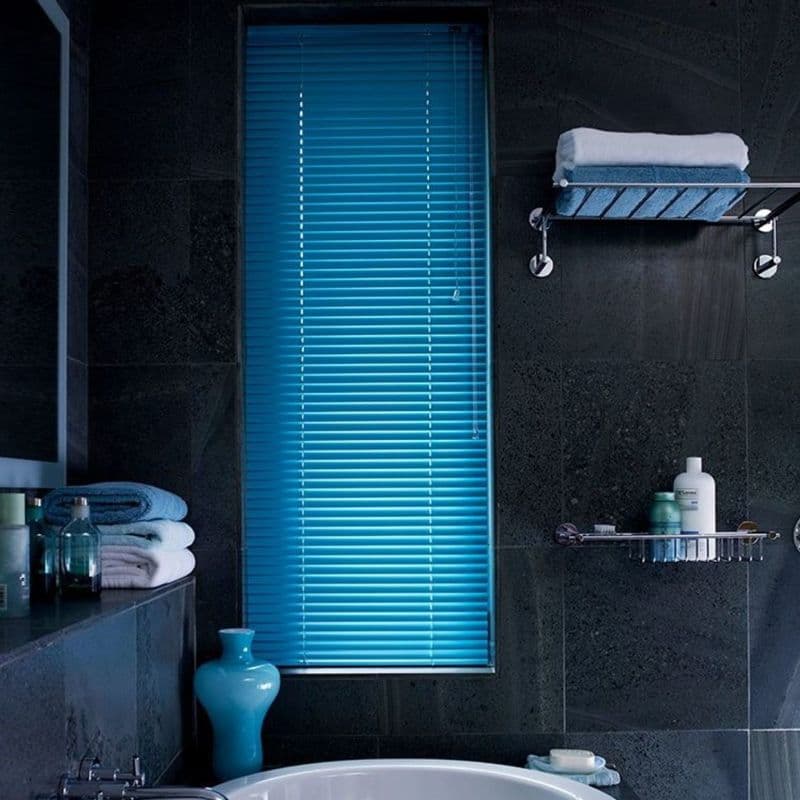 Blue horizontal Venetian blinds, closed, their slats letting narrow light strips through a narrow window in a dark slate-tiled bathroom with bathtub, folded towels, toiletries and a turquoise vase.