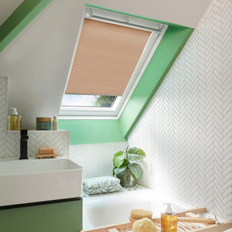 Beige pleated skylight blind, lowered halfway and filtering light, set within a mint-green dormer; surrounding attic bathroom shows white herringbone tiles, sink, bathtub, plant, and bath accessories.