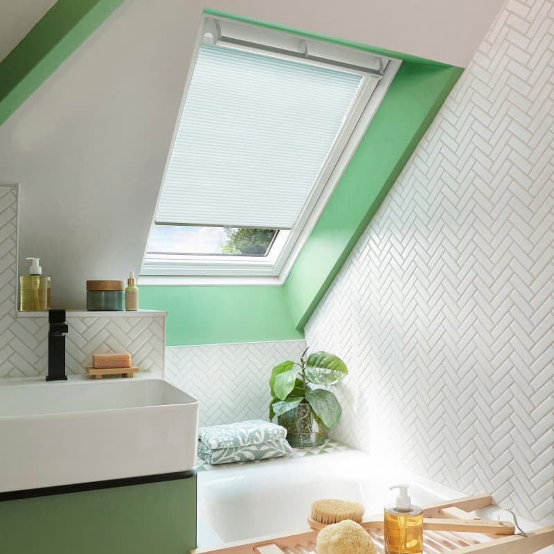 White pleated skylight blind, partially lowered across a slanted roof window, filtering soft daylight; bathroom with green-painted trim, white herringbone tiles, basin, bathtub ledge with plant and toiletries.