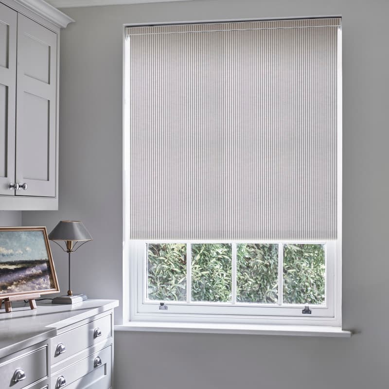 Striped pleated roller blind, lowered nearly to the sill with thin vertical pinstripes; blocking and softly diffusing daylight over a pale-grey room with white cabinetry, lamp, and leafy outside view.