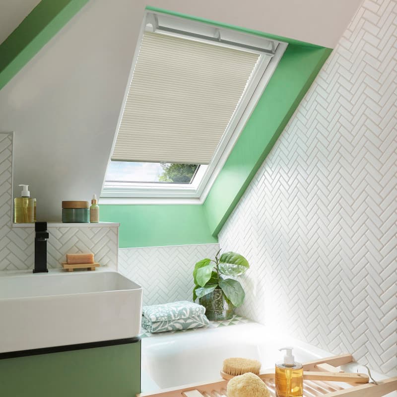 Cream pleated skylight blind, partially lowered and filtering daylight; set in a green-framed roof window above a white-tiled attic bathroom with sink, bathtub ledge, plant, towels and bath accessories.