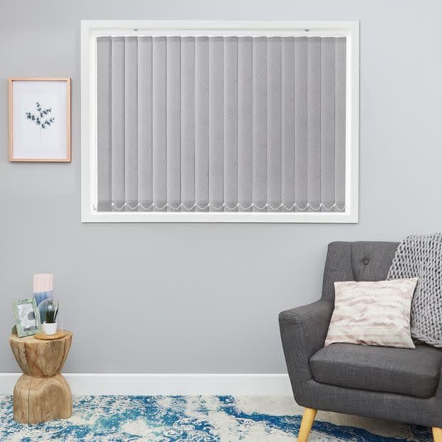 Vertical grey fabric blinds filling the window, with scalloped weighted hems; closed, softly diffusing daylight; in a small living room with a grey armchair, wooden side table and botanical print.