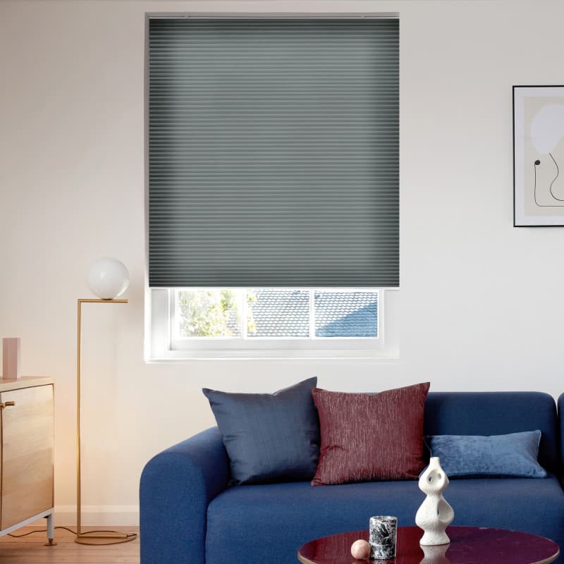 Dark grey pleated blind, lowered over most of the window leaving a narrow bottom gap; living room with blue sofa, cushions, floor lamp, and roof tiles and trees visible beyond.