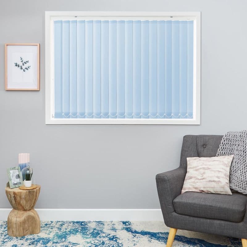 Light-blue vertical blinds covering a rectangular window, closed and gently filtering daylight; in a gray living room with framed botanical art, wooden side table, patterned blue rug, and gray armchair.