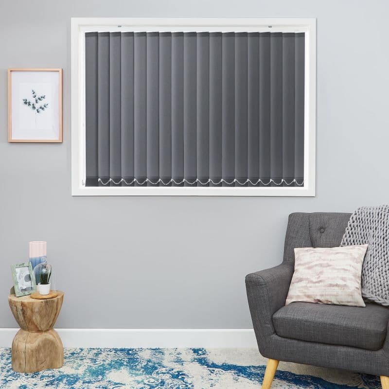Dark grey vertical blinds, fully closed with a bottom bead chain linking slats, covering a white-framed window; in a lit living room with grey armchair, wooden side table and blue rug.