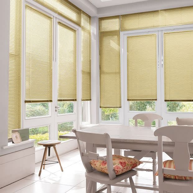 Beige pleated Venetian-style blinds cover tall French windows and doors, lowered and filtering soft daylight; in a bright tiled dining area with white table, chairs, floral cushions and garden view.