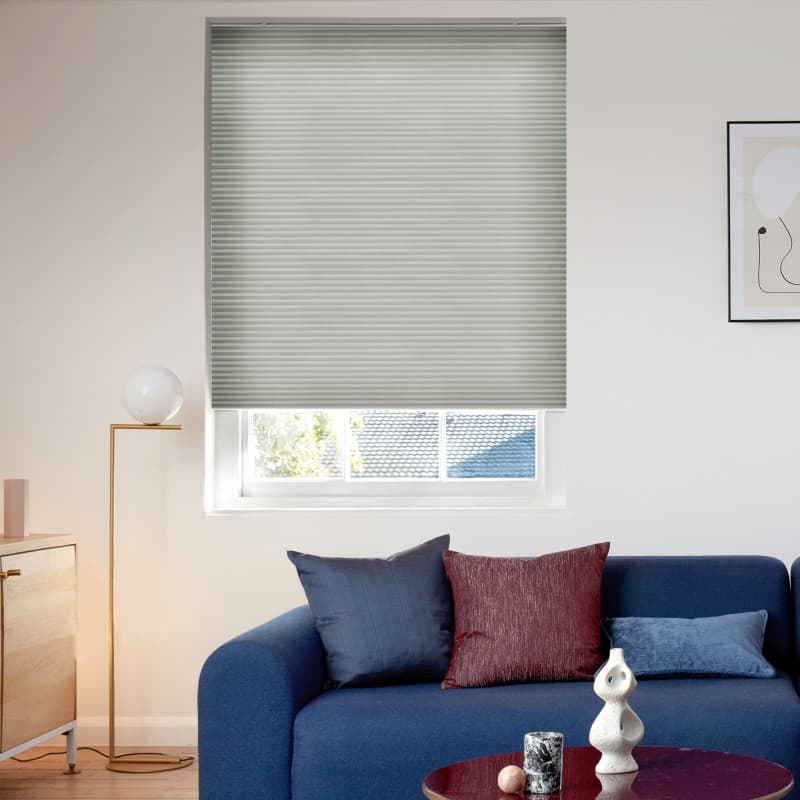 Light-gray cellular blind, horizontally pleated, fully lowered over a window, filtering daylight; in a sunlit living room with a blue sofa, assorted cushions, a floor lamp and small coffee table.