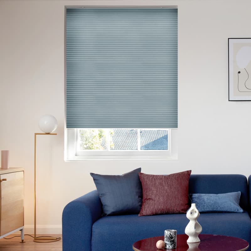 Light-blue pleated cellular blind, lowered to just above the sill leaving a bottom gap, filtering daylight over a modern living room with a blue sofa, cushions, lamp and wall art.