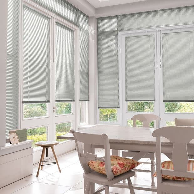 Light-grey Venetian blinds on tall windows and glass doors, lowered to mid-height and partially closed, filtering daylight into a dining area with white table, chairs, floral cushions, garden view.