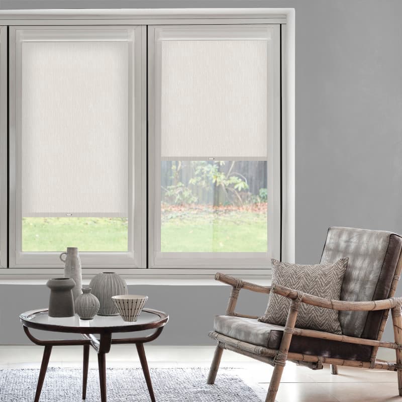 White roller blinds partially lowered over two windows, filtering daylight; gray living room with low coffee table holding ceramics and a bamboo-rattan armchair facing a grassy backyard.
