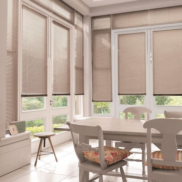 Beige horizontal blinds covering tall French doors and windows, lowered with slats mostly closed and softly filtering daylight; sunlit dining corner with wooden table, chairs, floral cushions and garden view.