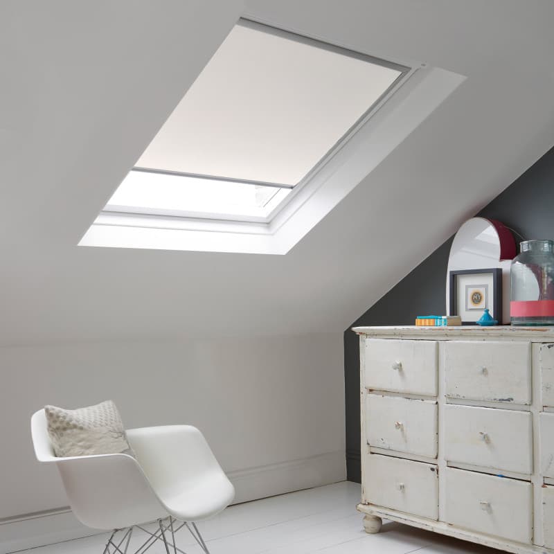Room with white eaves wall, dark blue-grey wall to right with a shabby chic white drawer unit, a contemporary white chair, white painted floorboards and a skylight blind in lily white.