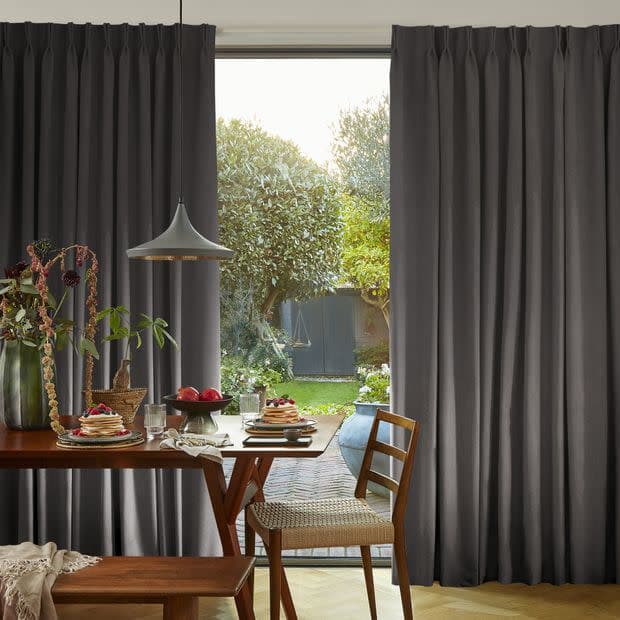 Floor-length pleated dark-gray curtains, parted at center, framing a door; they reveal a sunlit backyard with trees and lawn beyond a wooden dining table set with dishes and pendant lamp.