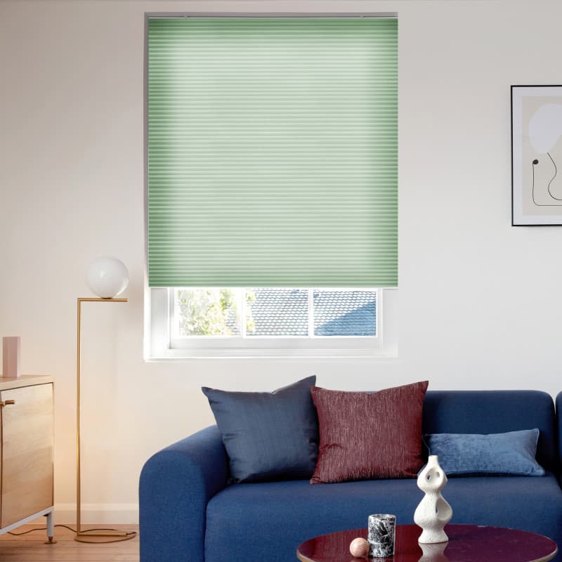 Mint-green pleated cellular blind lowered to cover most of the window, softly filtering daylight and partially obscuring roof and trees; modern living room with blue sofa, cushions, lamp and table.