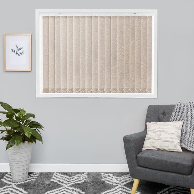 Beige patterned vertical blinds, scalloped bottoms, fitted inside a white frame â€” closed, softly filtering daylight. Context: grey-walled living room with armchair, cushion, potted plant and framed print.