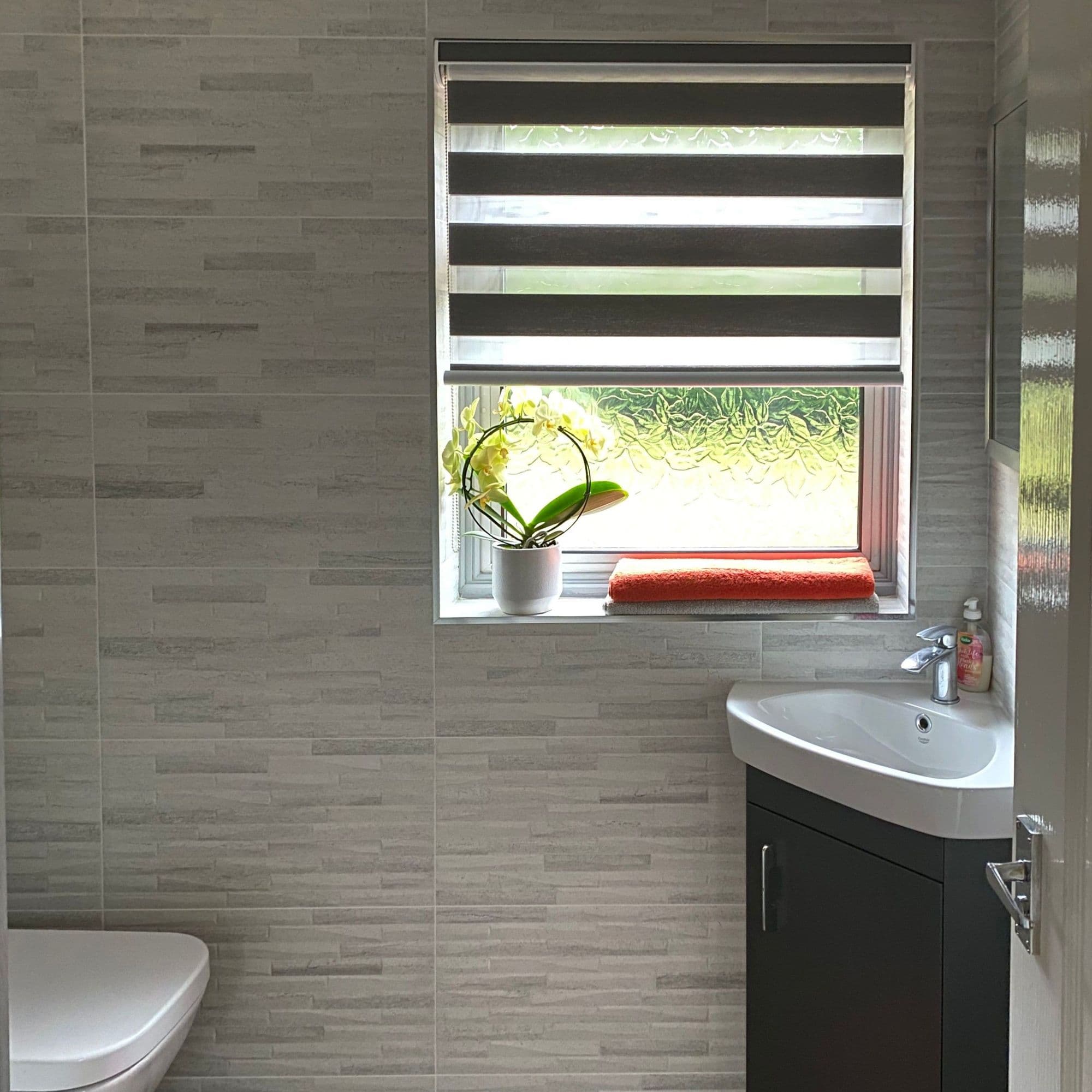 Dual-layer zebra roller blind, horizontal opaque-and-sheer stripes, partially lowered, filtering daylight; frosted sill holds potted orchid and folded orange towel in tiled bathroom with corner sink, toilet.