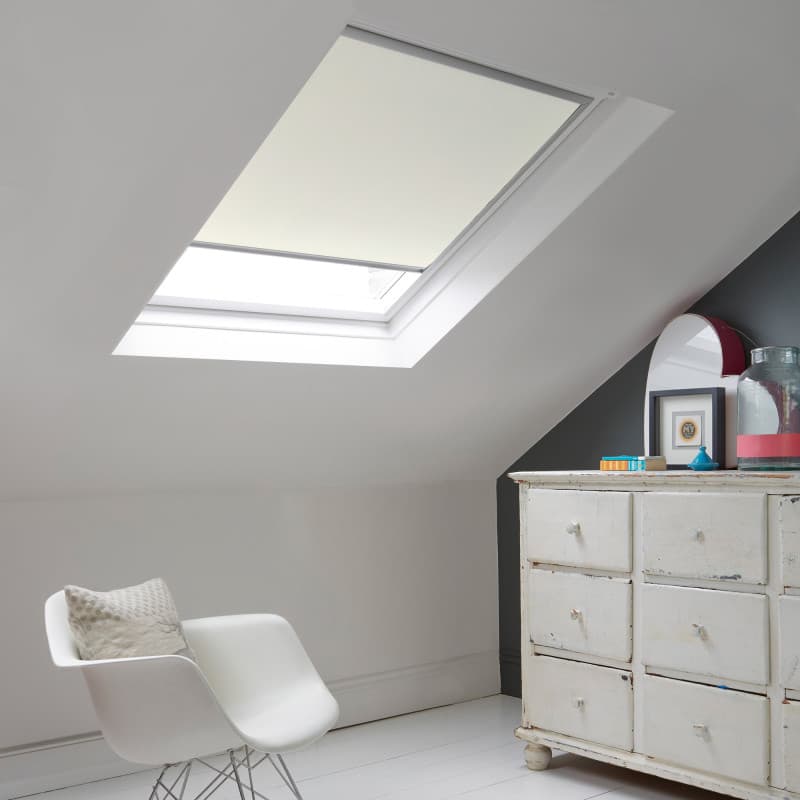 Room with white eaves wall, dark blue-grey wall to right with a shabby chic white drawer unit, a contemporary white chair, white painted floorboards and a skylight blind in pale cream.