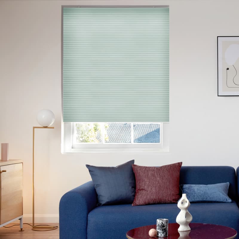 Pale mint cellular blind, fully lowered with subtle horizontal pleats, filtering daylight, above a partially visible window in a bright modern living room with a blue sofa, cushions, floor lamp.