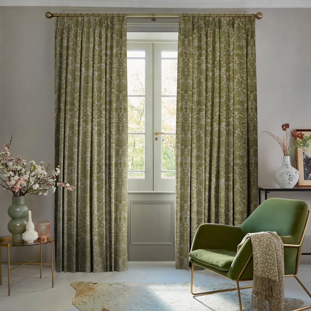 Patterned green pleated curtains on a brass rod, drawn slightly apart revealing French doors; filtering soft daylight; in a neutral living room with a green chair and floral accents.