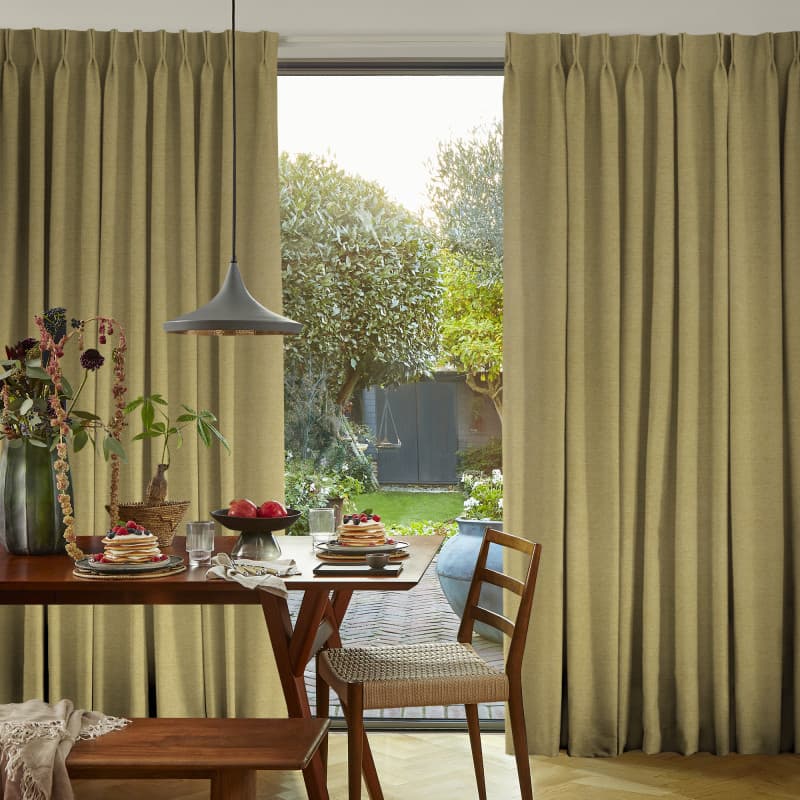 Beige floor-length pleated curtains, slightly parted, framing and filtering sunlight through a sliding glass door; dining table set with pancakes, pendant lamp and wooden chairs, garden view beyond.