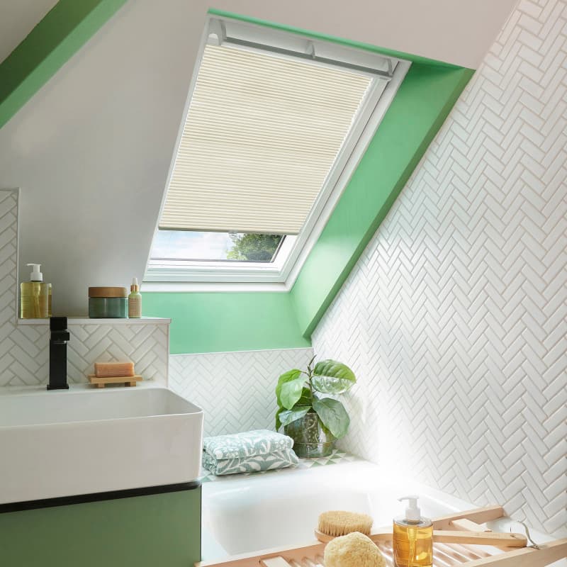 Cream pleated skylight blind, lowered halfway and filtering daylight, in a bright sloped-ceiling bathroom with green trim, white herringbone tiles, sink, bathtub and potted plant.