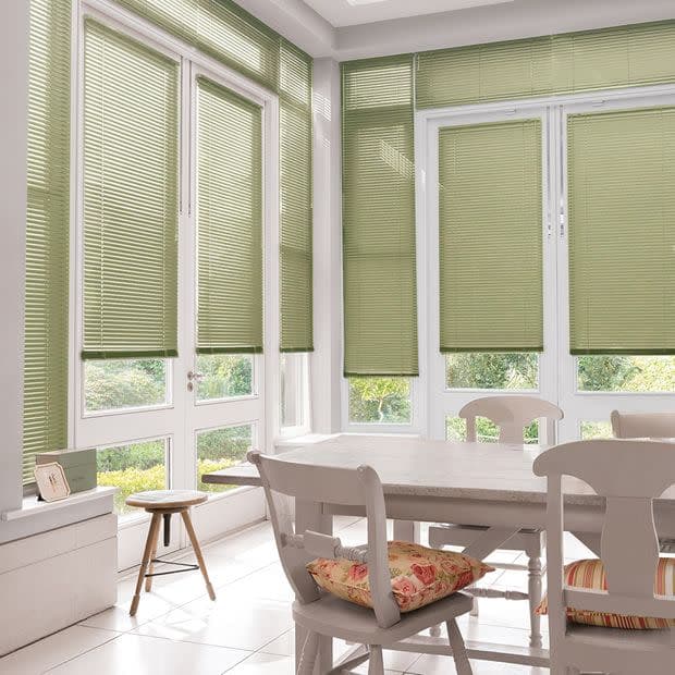 Green pleated window blinds, fully lowered across tall French doors and surrounding windows, lightly filtering sunlight; dining room with white wooden table and chairs, floral cushions, tile floor, garden view.